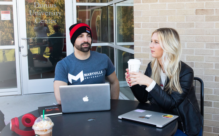Two Maryville students sitting outside at a table drinking coffee and using laptops